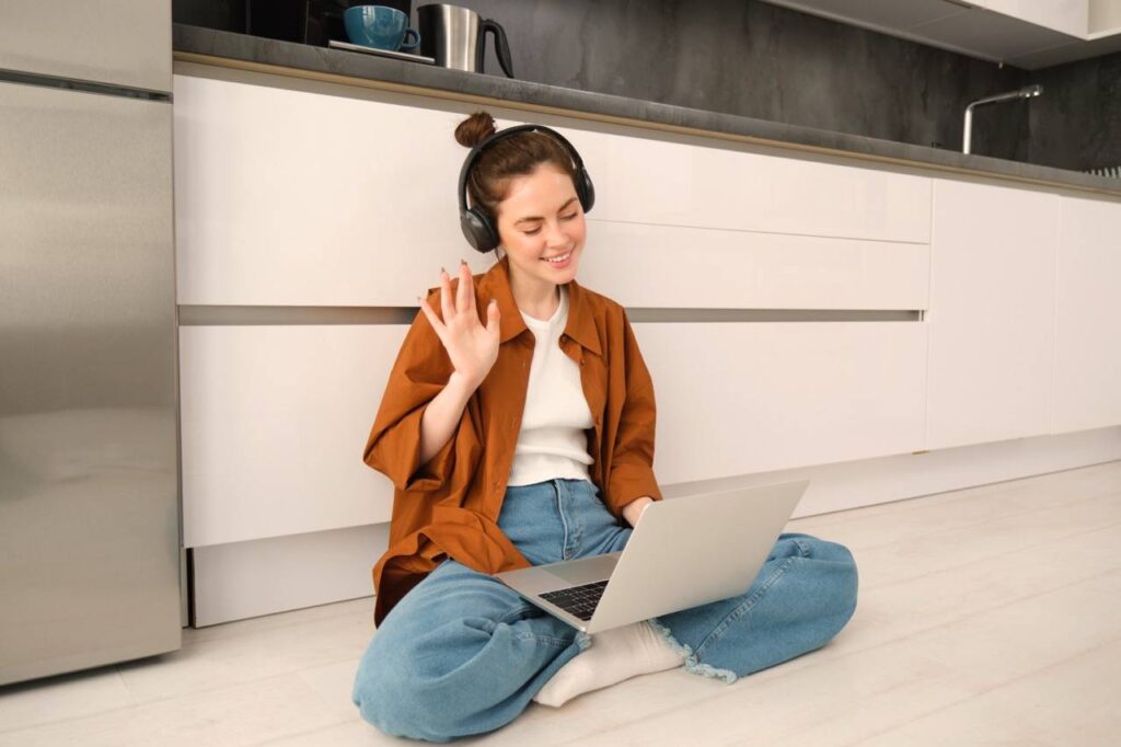 Portrait of happy woman in headphones, looking at laptop, waving hand at computer, chatting on video online, joins team meeting, calling friend on computer, sitting on floor